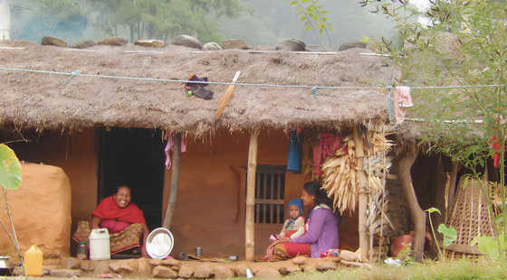 Maison traditionnelle en torchis avec toit de chaume, où une famille népalaise s’affaire devant l’entrée.