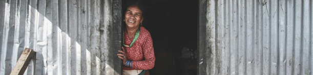 Femme népalaise souriante à l’entrée de sa maison en tôle, baignée par la lumière du matin.