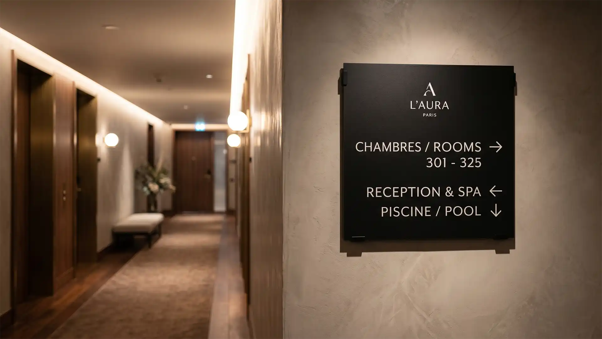 Un panneau de signalisation moderne noir mat fixé au mur dans un couloir d'hôtel de luxe. Le panneau affiche le logo "L'AURA PARIS" et des directions en blanc : "CHAMBRES / ROOMS 301 - 325" avec une flèche vers la droite, "RECEPTION & SPA" avec une flèche 