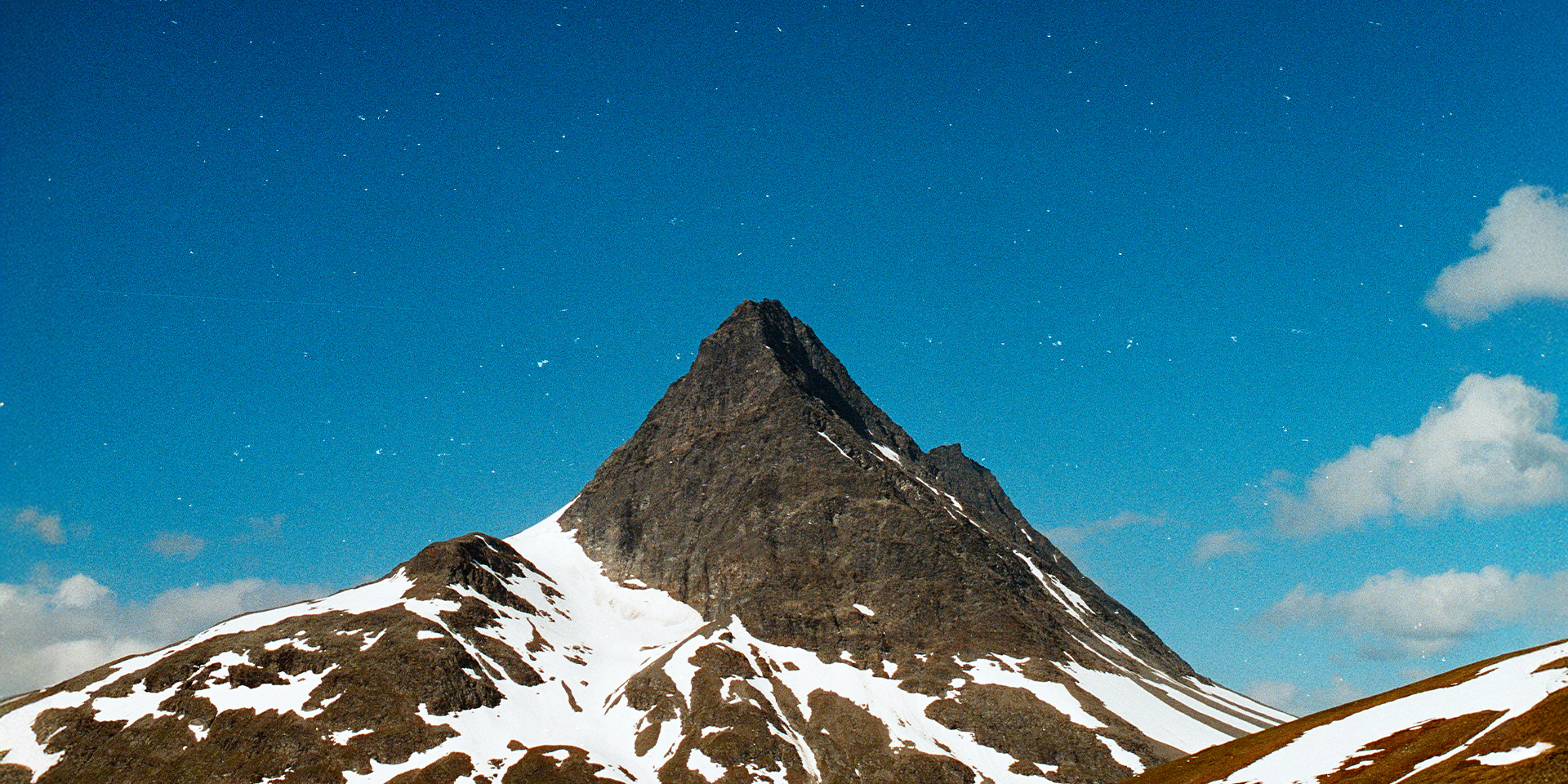 Snow-capped mountain peak in Norway under a clear blue sky with scattered clouds. Analog. 