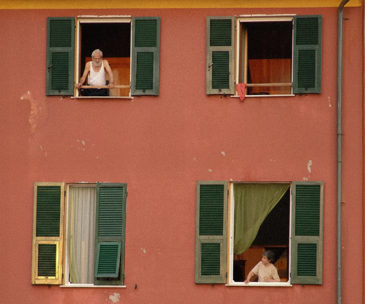 Older man and woman looking out from separate open windows with green shutters on a weathered red building facade.