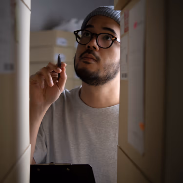 Mann mit Brille und Mütze, der zwischen Regalen mit Aktenordnern nach etwas schaut und einen Stift hält.
