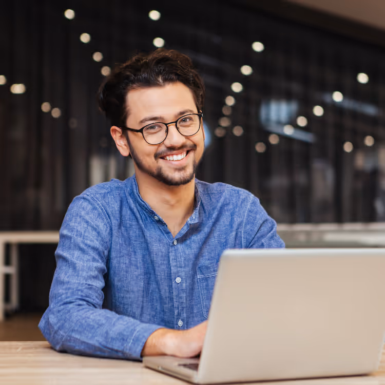 Lächelnder Mann mit Brille und blauem Hemd sitzt an einem Schreibtisch vor einem Laptop.