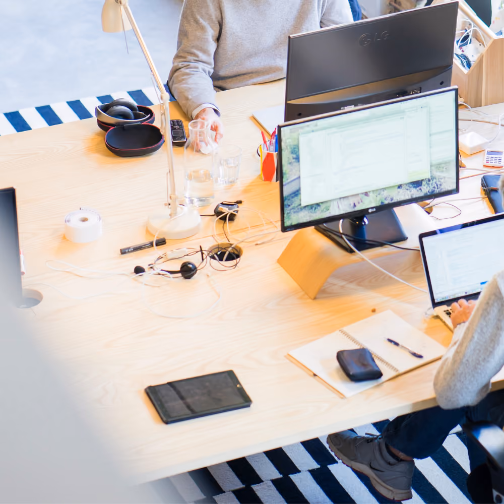 Zwei Personen arbeiten an einem Tisch mit Computermonitoren, Laptop, Notizbuch, Kopfhörern und einer Tischlampe.