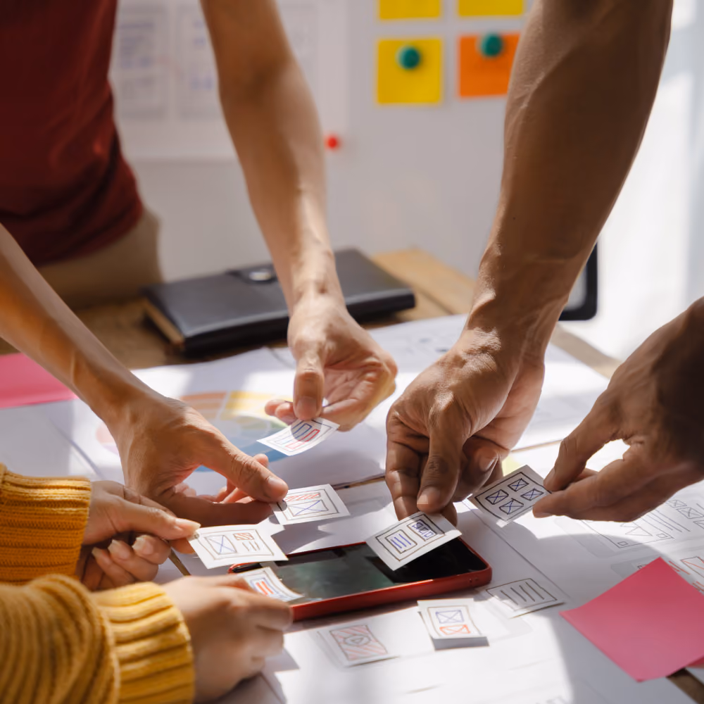 Menschen arbeiten gemeinsam an einem Design-Layout mit kleinen Papierstücken und einem Smartphone auf einem Tisch.