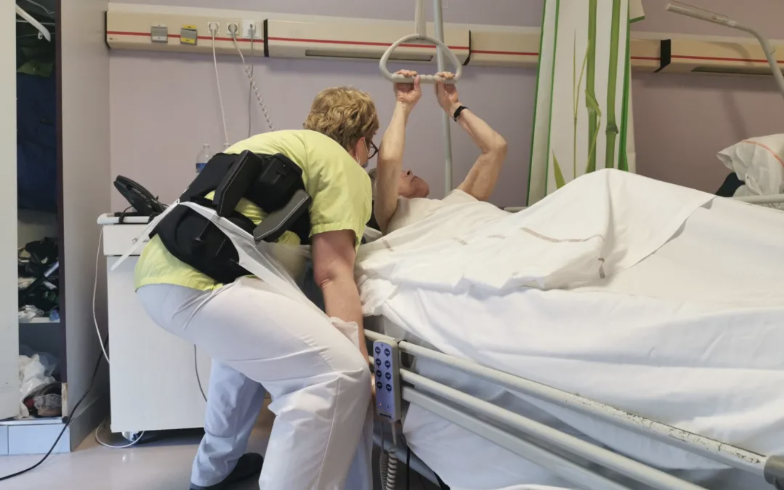 Une infirmière aide une patiente à se relever. Pour cela elle porte un exosquelette pour protéger son dos