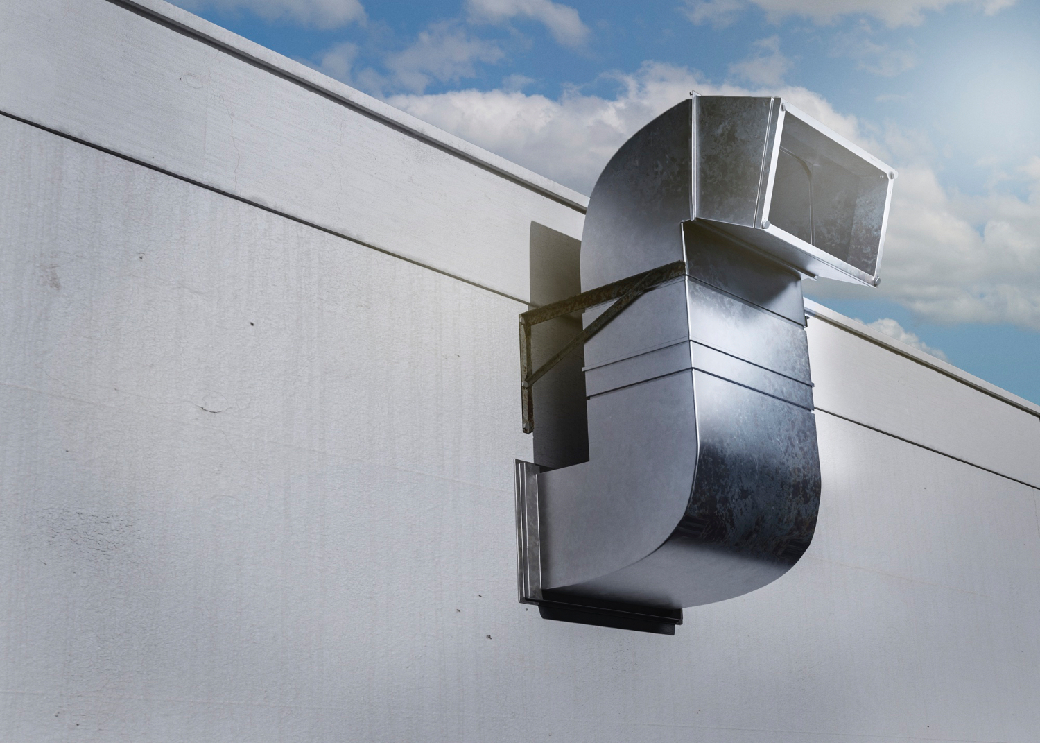 Metal ductwork mounted on a building wall against a blue sky and clouds.