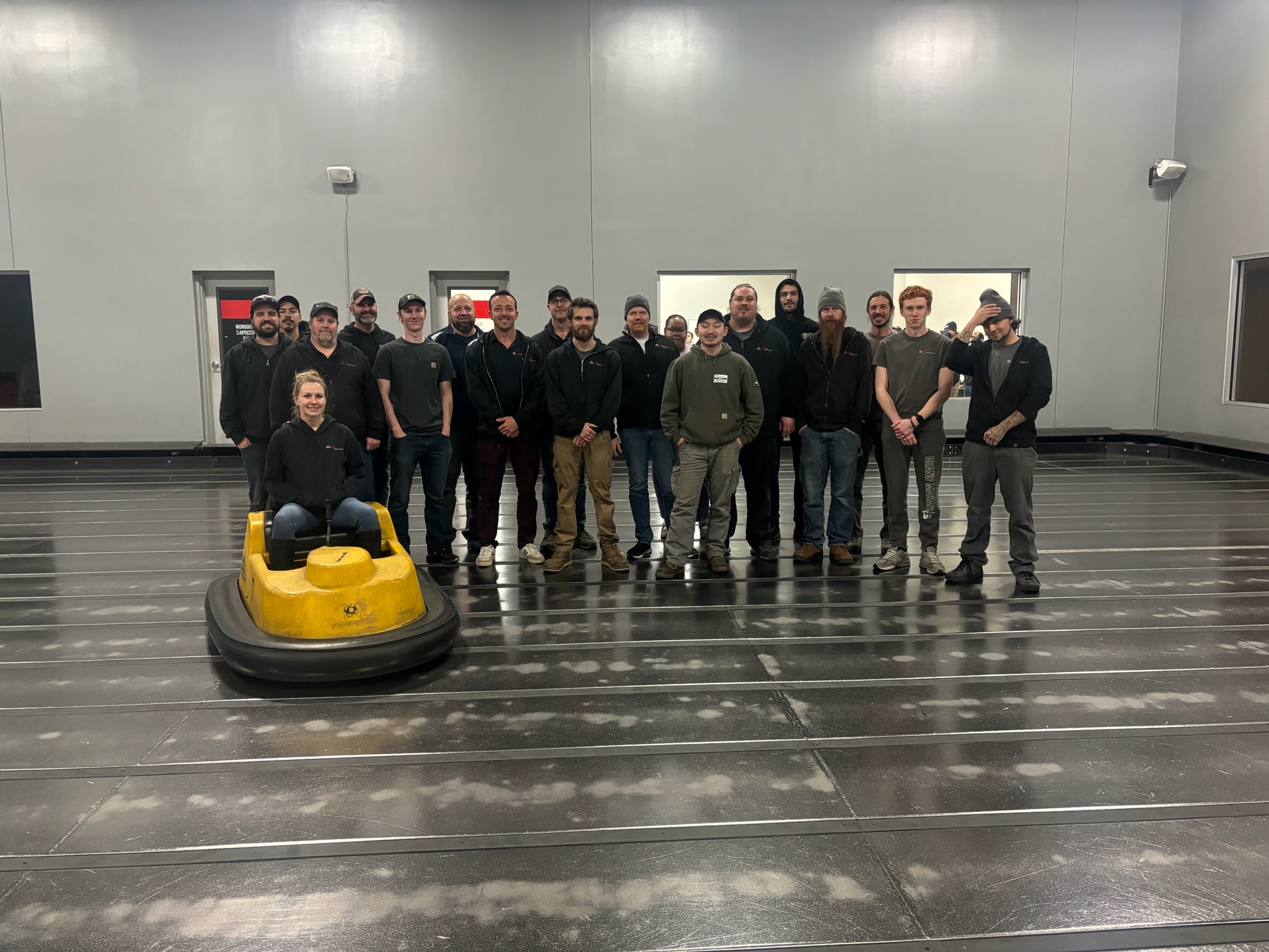 A team of employees poses together with a yellow bumper car in a spacious facility.
