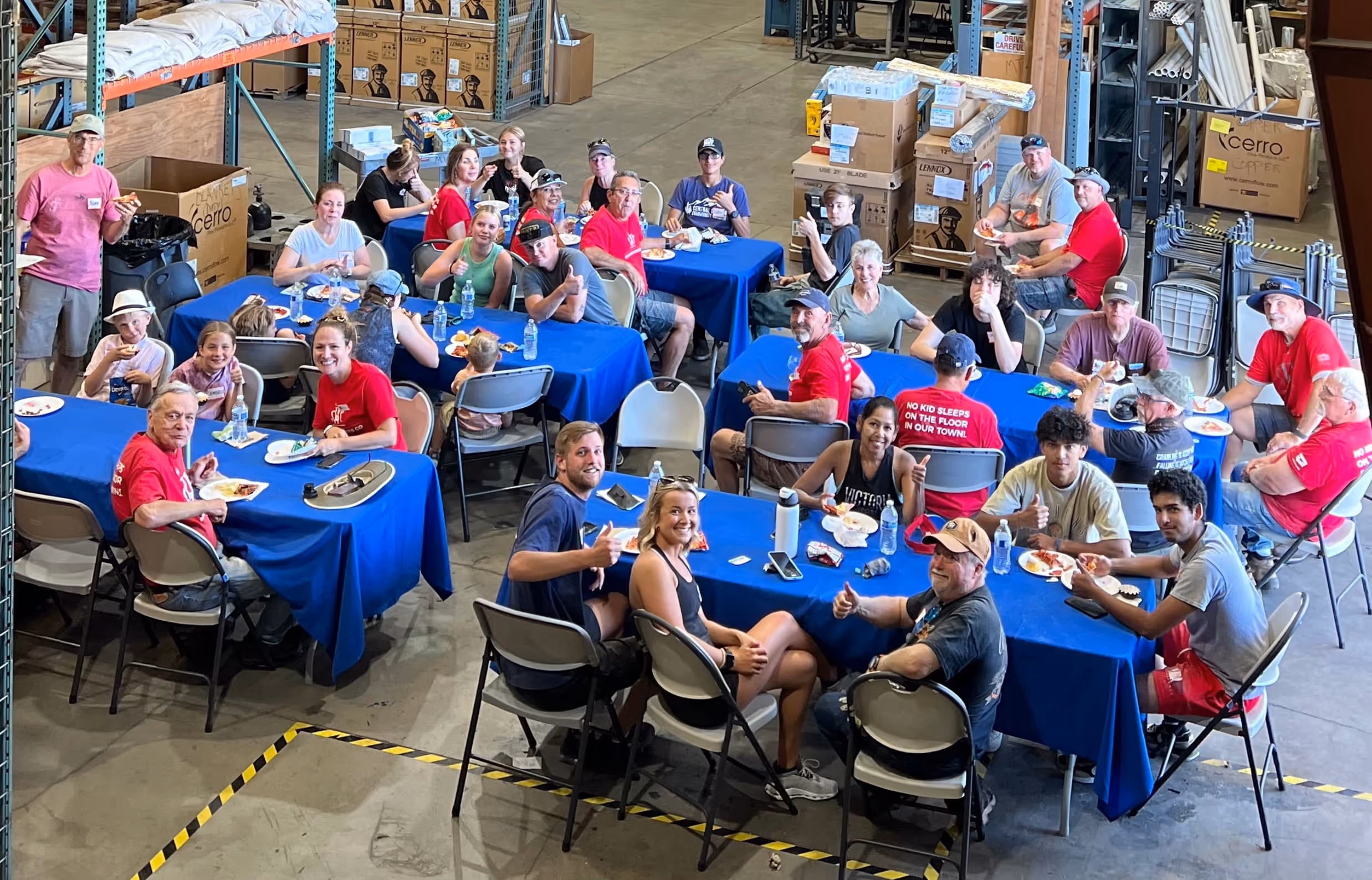A group of people sits around tables in a warehouse, enjoying a meal together.