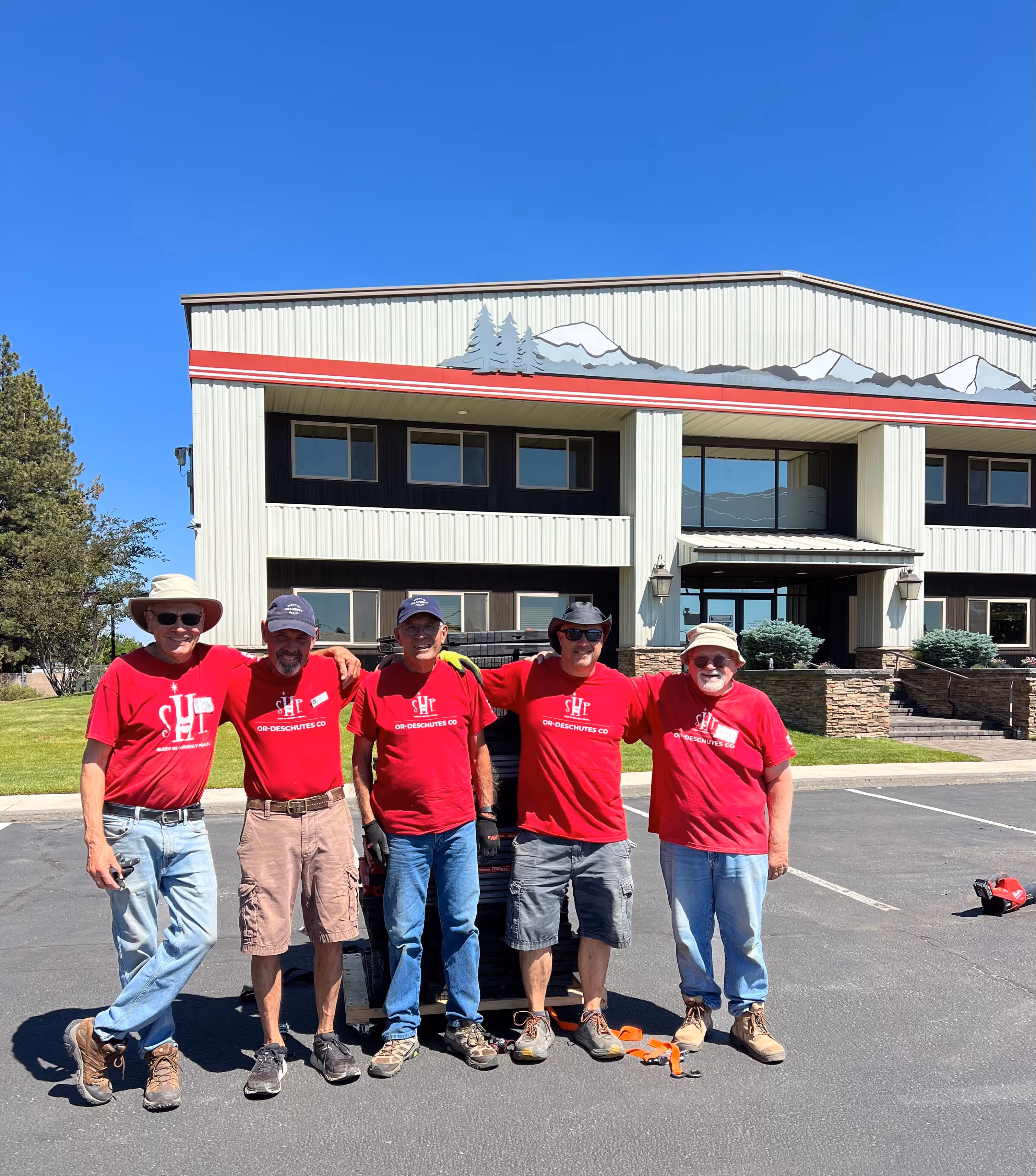 Five smiling workers in red shirts stand together in front of a building at a construction site.
