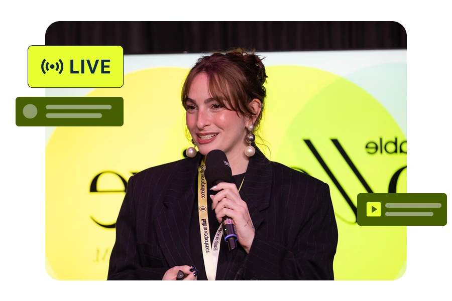 A woman with pearl earrings and a black pinstripe blazer holds a microphone and smiles while speaking at an event with a yellow and green background.