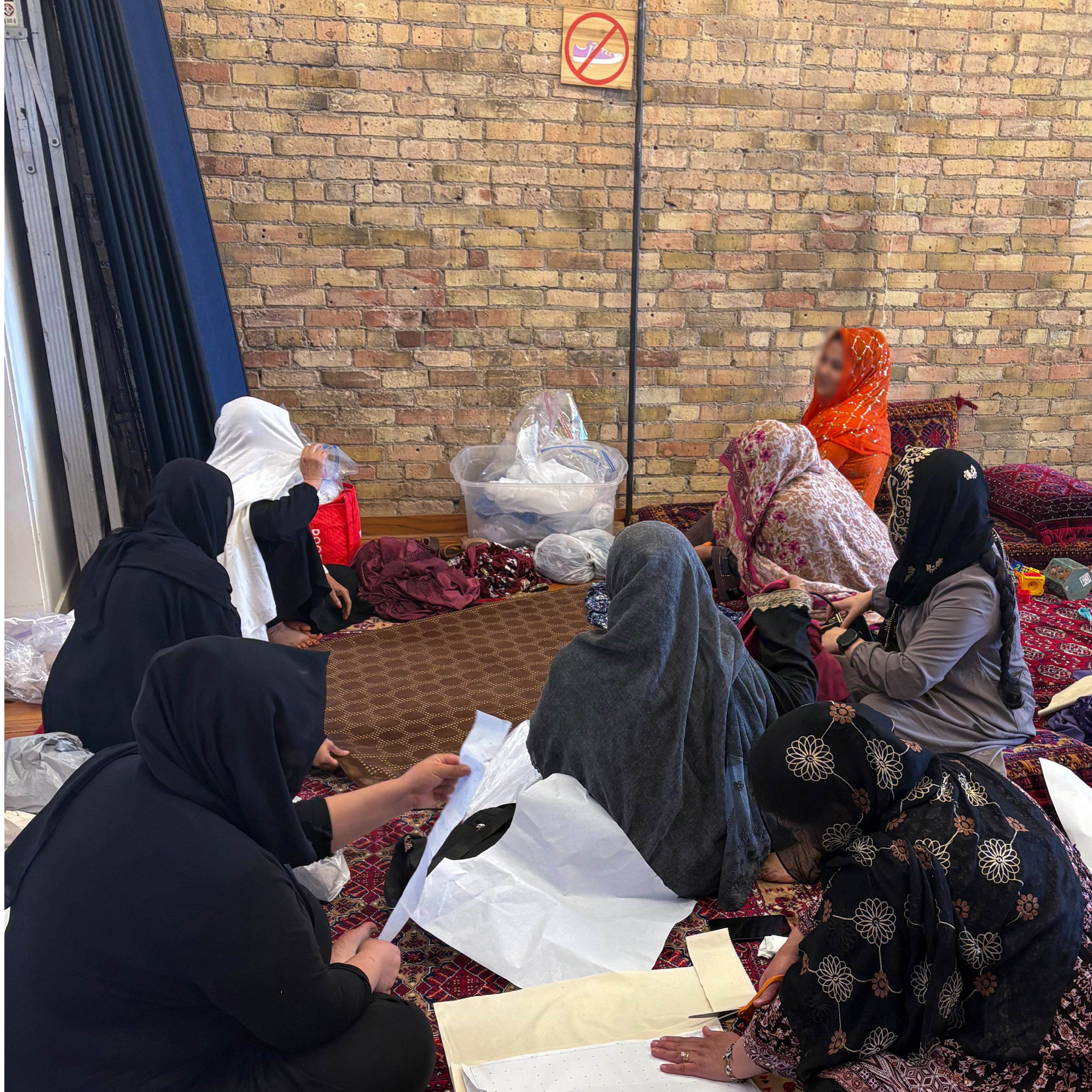 A group of Afghan women seated on the floor, some of them are examining textiles or paper sewing patterns.