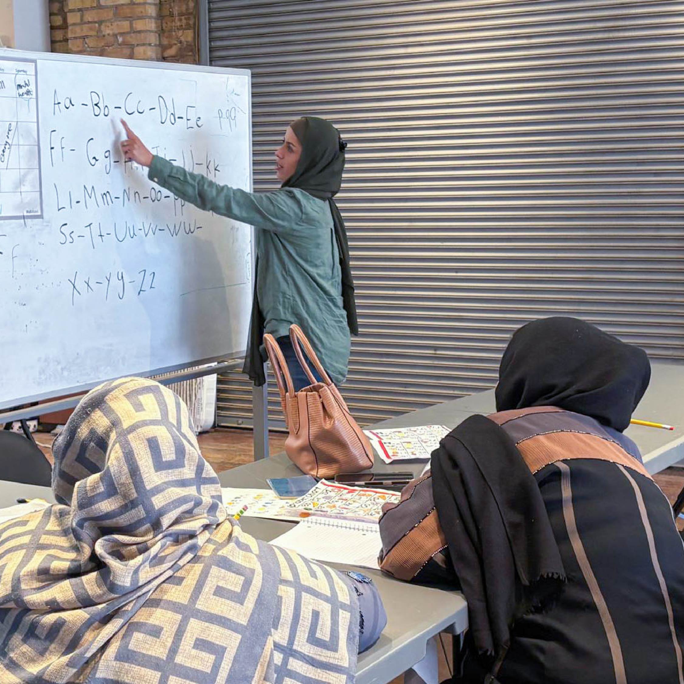Two women are watching an instructor review the English Alphabet on a whiteboard.