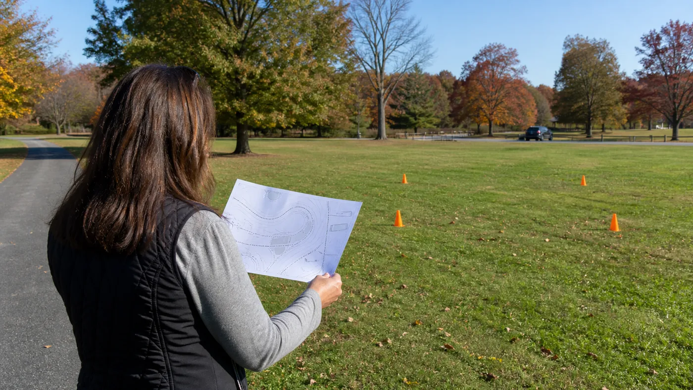 Event organizer reviewing a printed park map beside a paved path and a lawn marked with small cones.