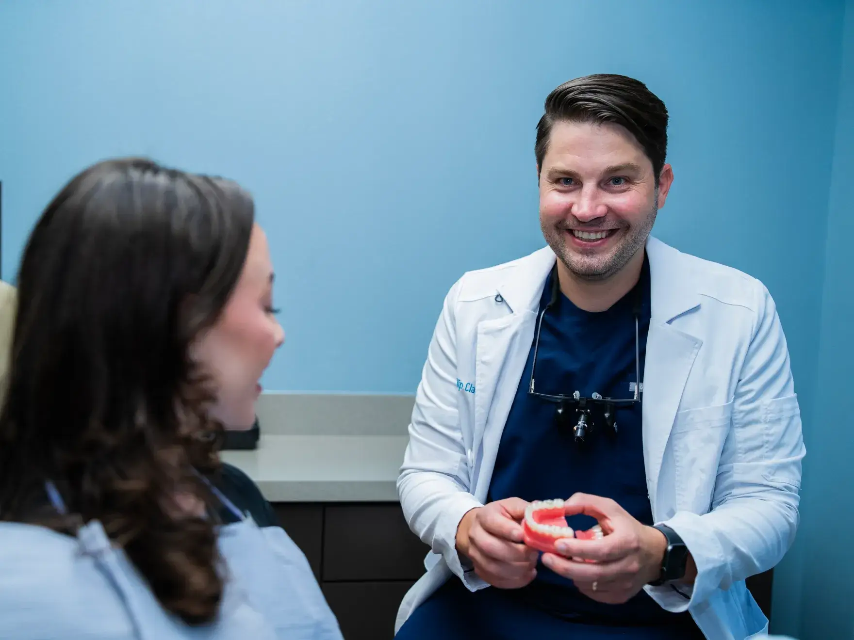 A dentist wearing magnifying glasses smiles while showing a dental tool to a patient in a chair.