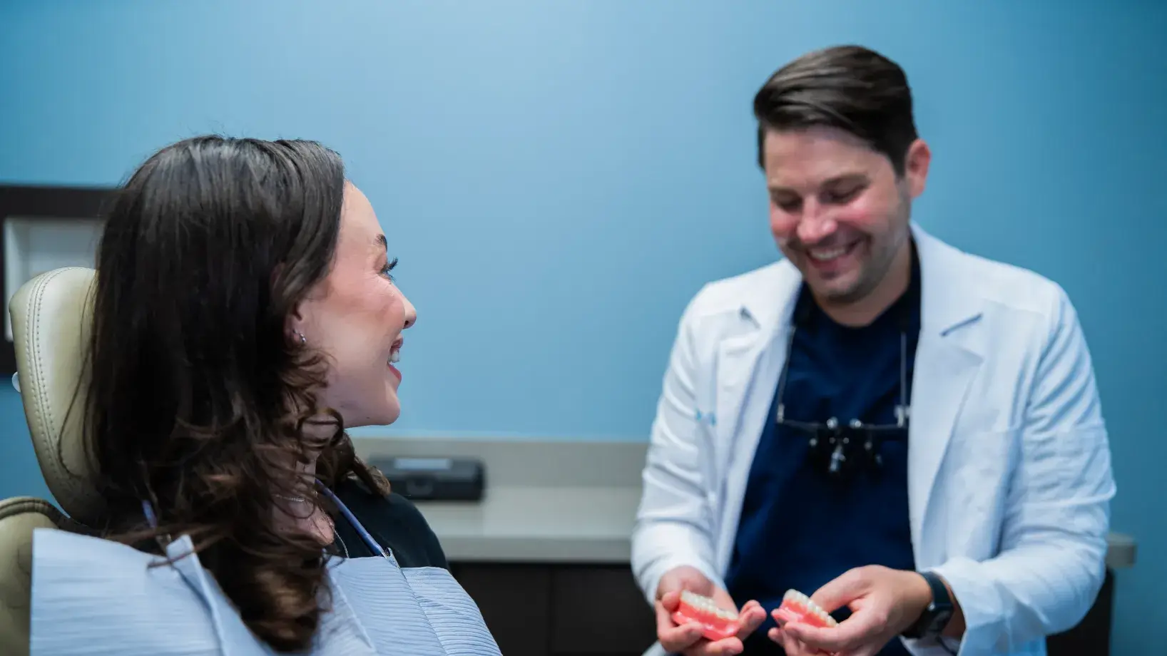 A smiling dental hygienist in a blue uniform holding dental tools in a dental office.