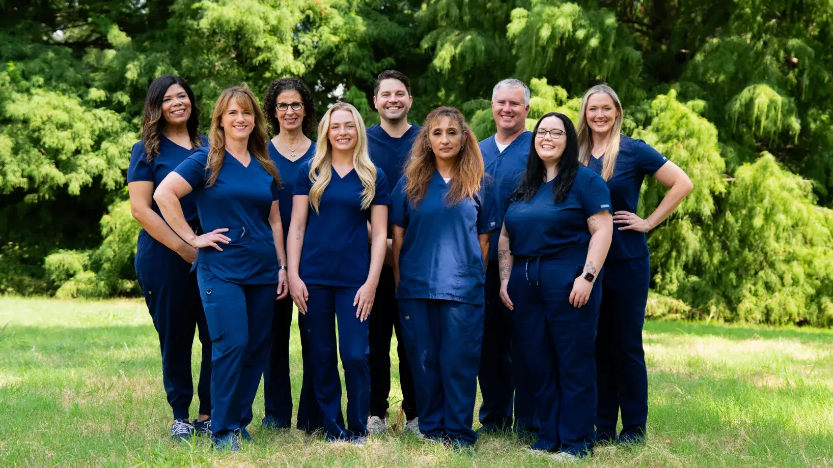 A group of nine people in blue scrubs standing outside on grass, smiling at the camera.