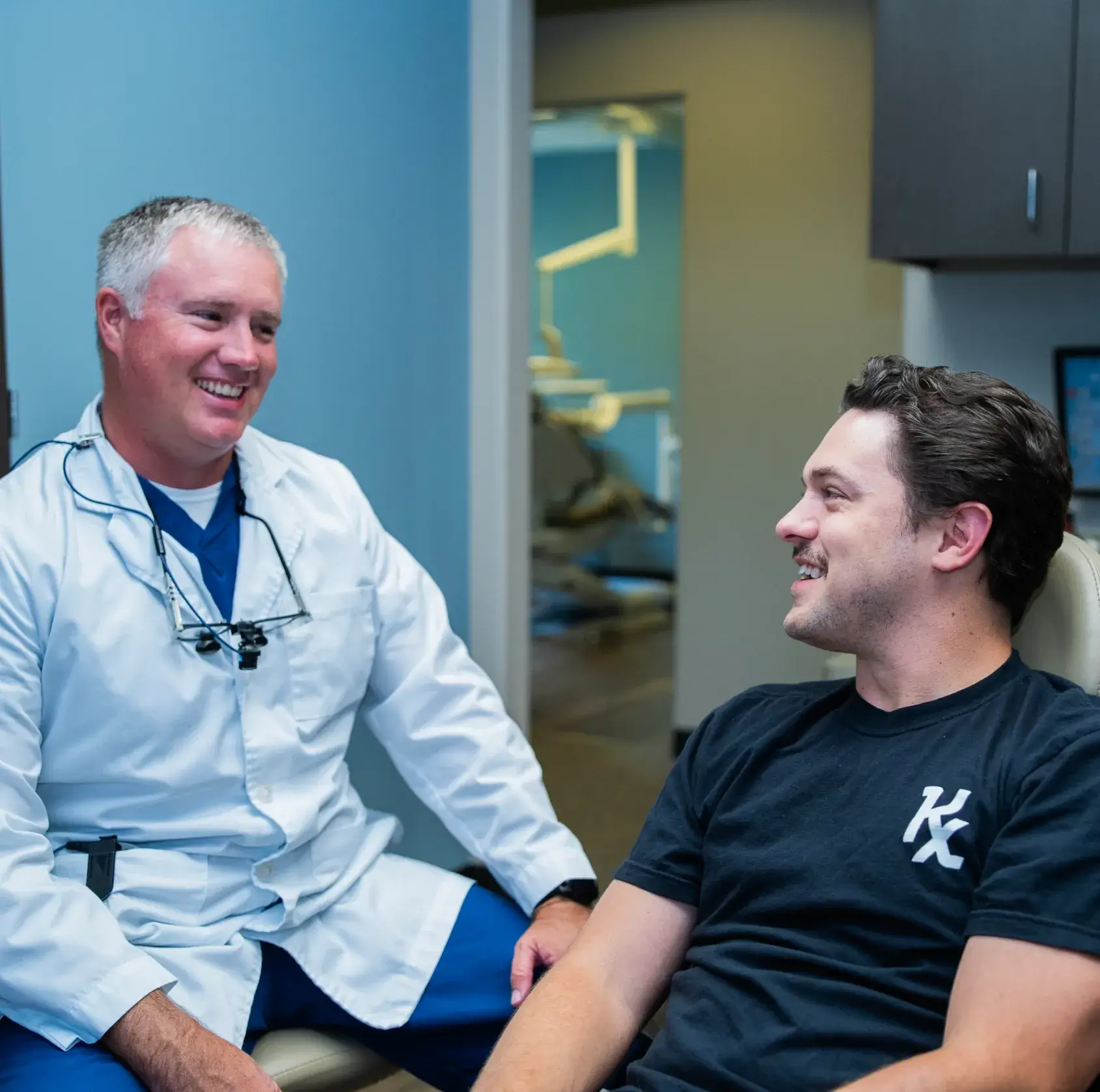 A dentist talks and smiles with a seated patient in a dental office.