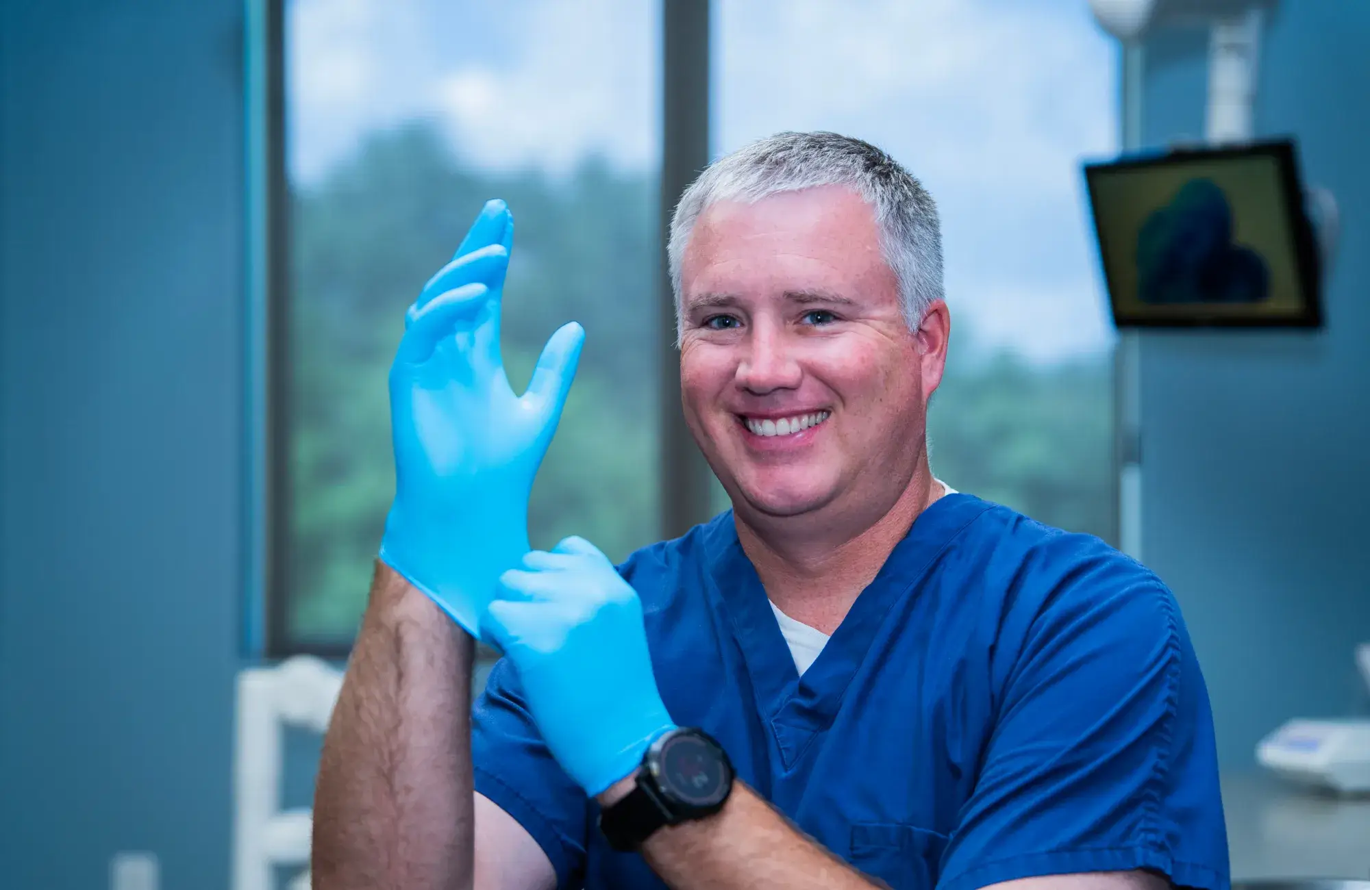 A smiling person in blue scrubs puts on blue gloves, standing indoors near a window.