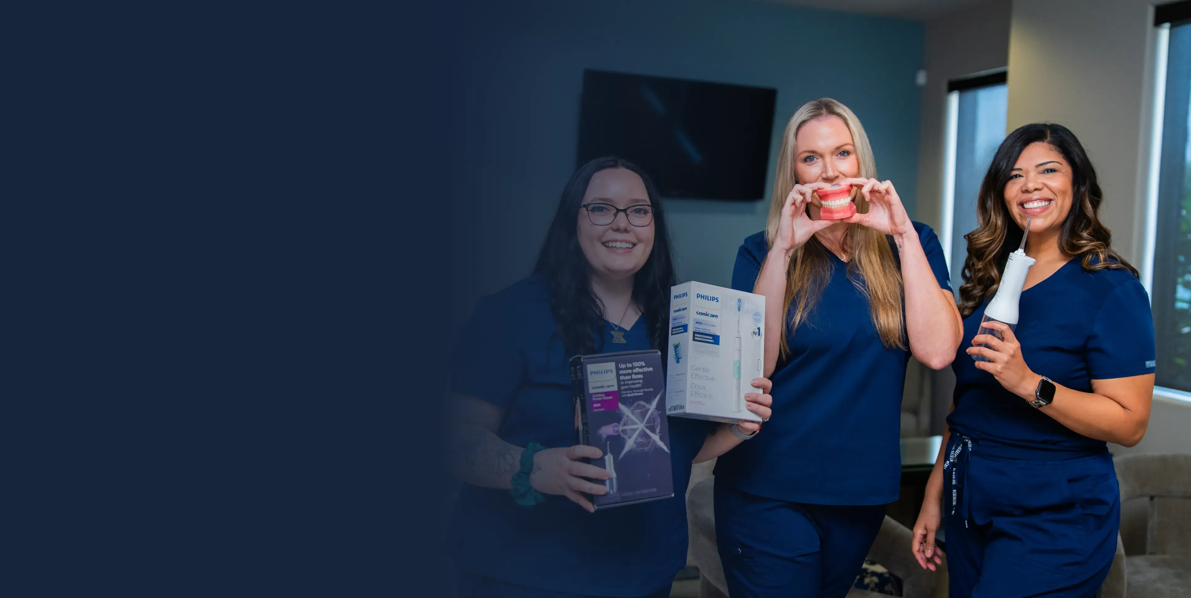Three people in blue uniforms smiling, holding dental care products and a dental model.