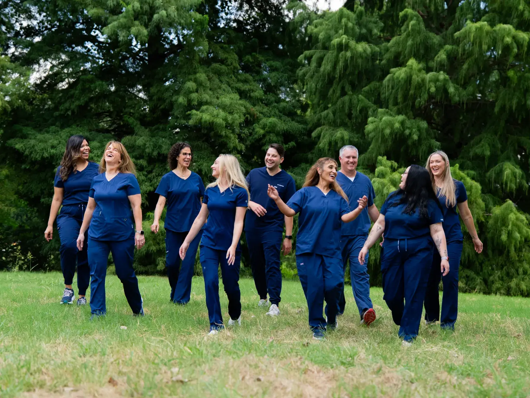 A group of nine people in blue uniforms walk and smile together on grass.