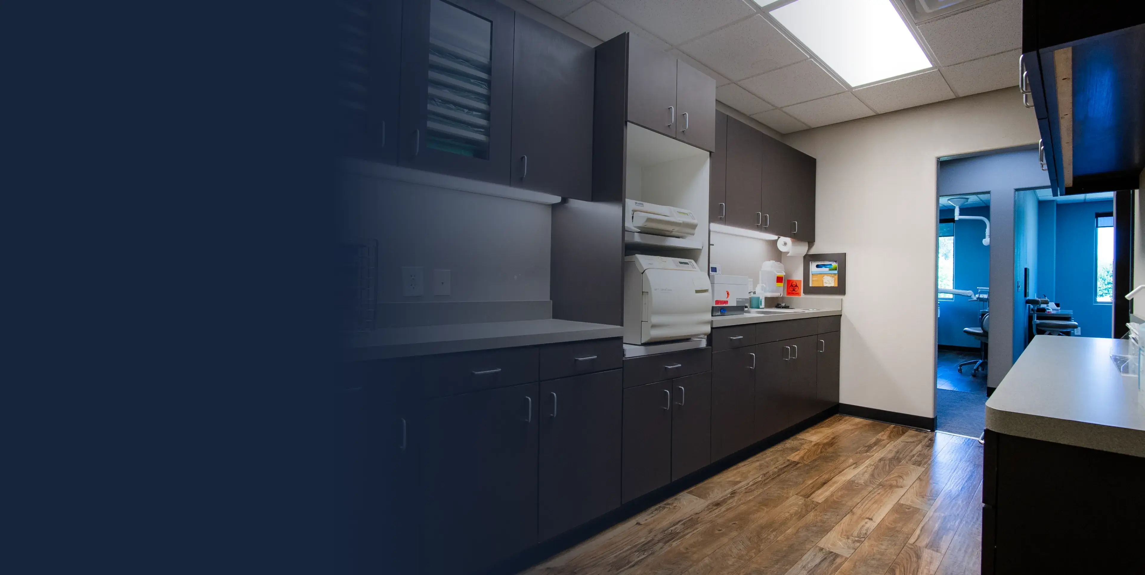 A dentist's office room with wooden floors, dark cabinets, and a sterilizer machine on the counter.