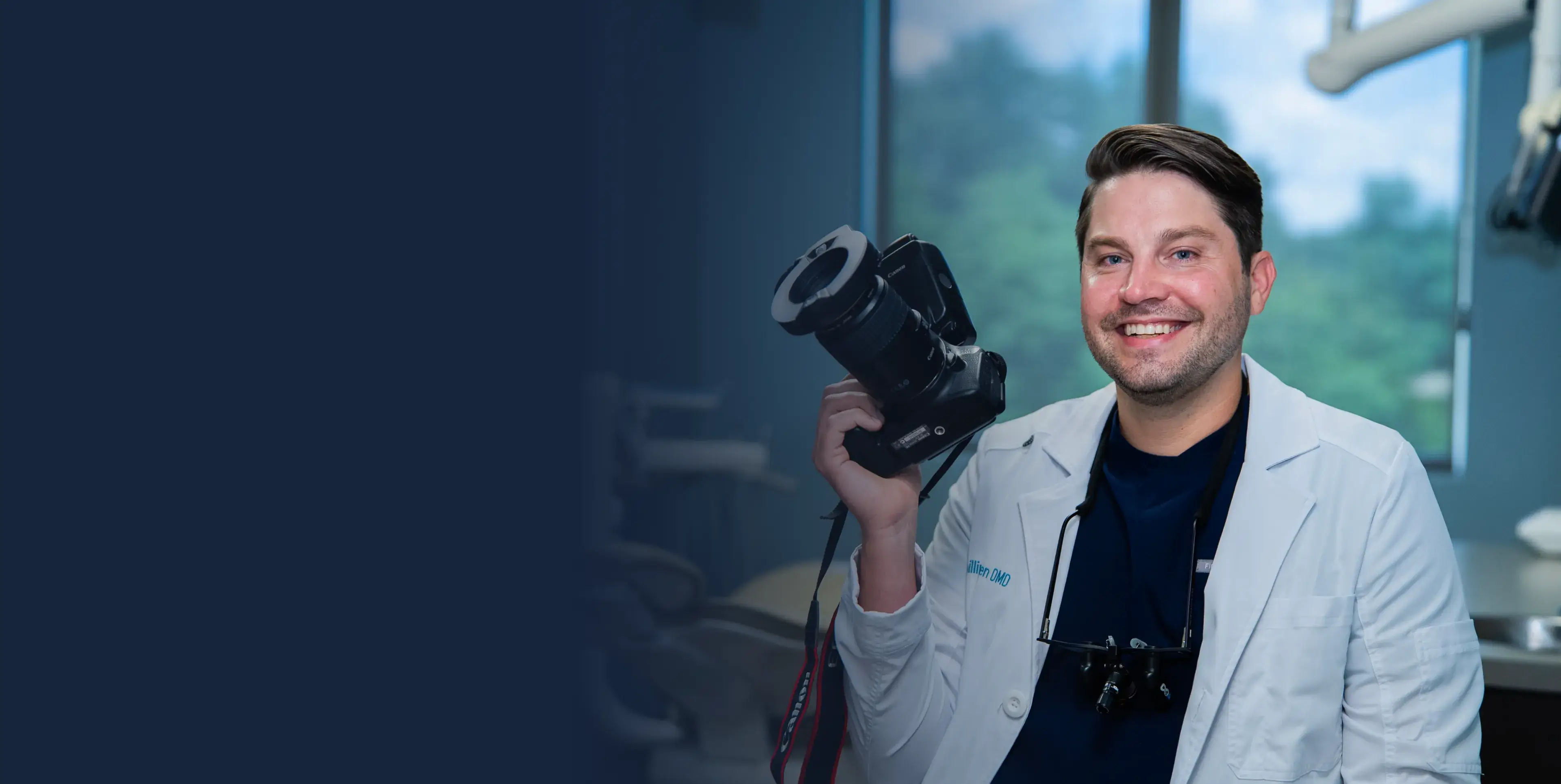 Smiling man in a white coat holding a camera, sitting in front of a large window.