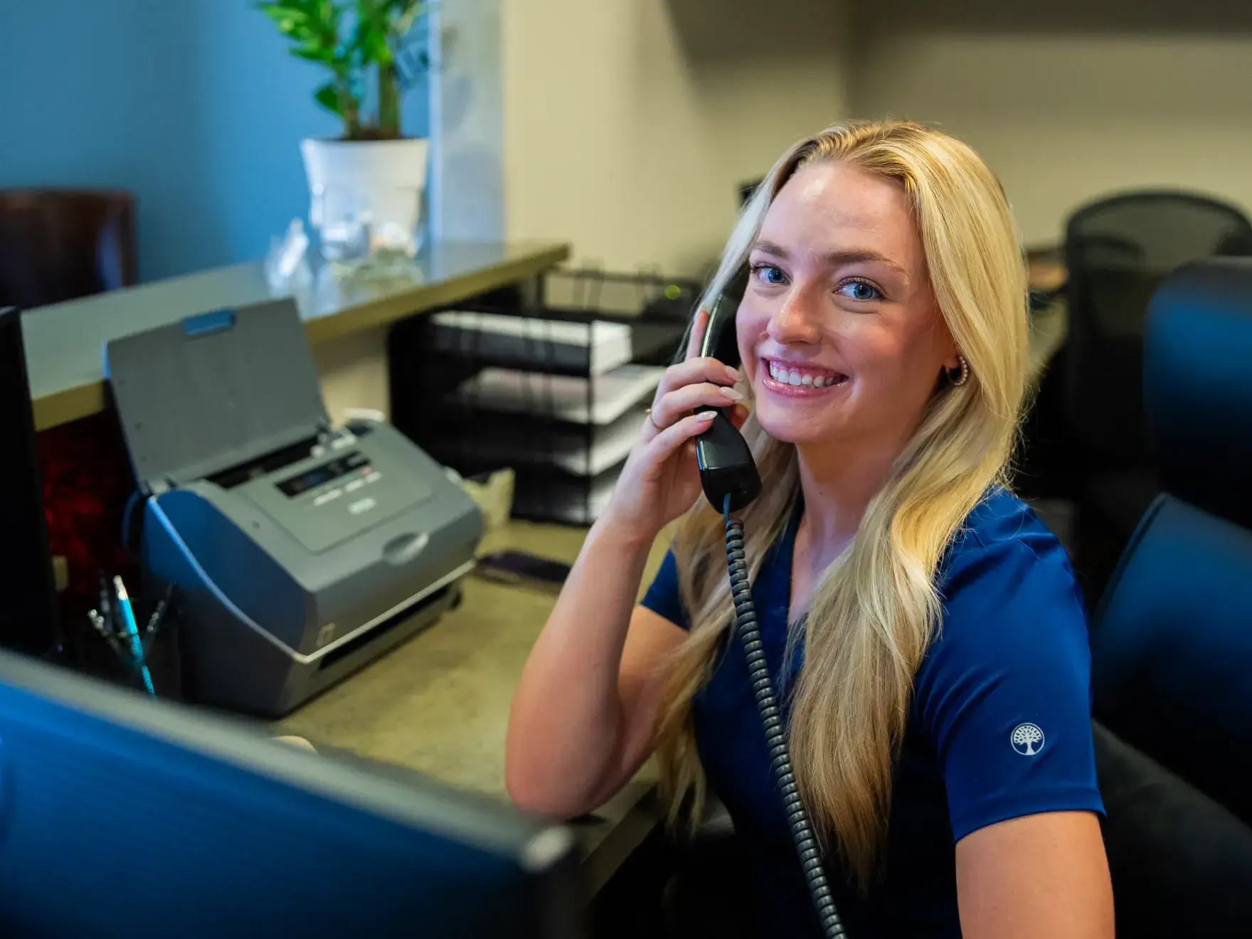 A woman in a blue shirt sits at a desk, smiling and talking on a phone.