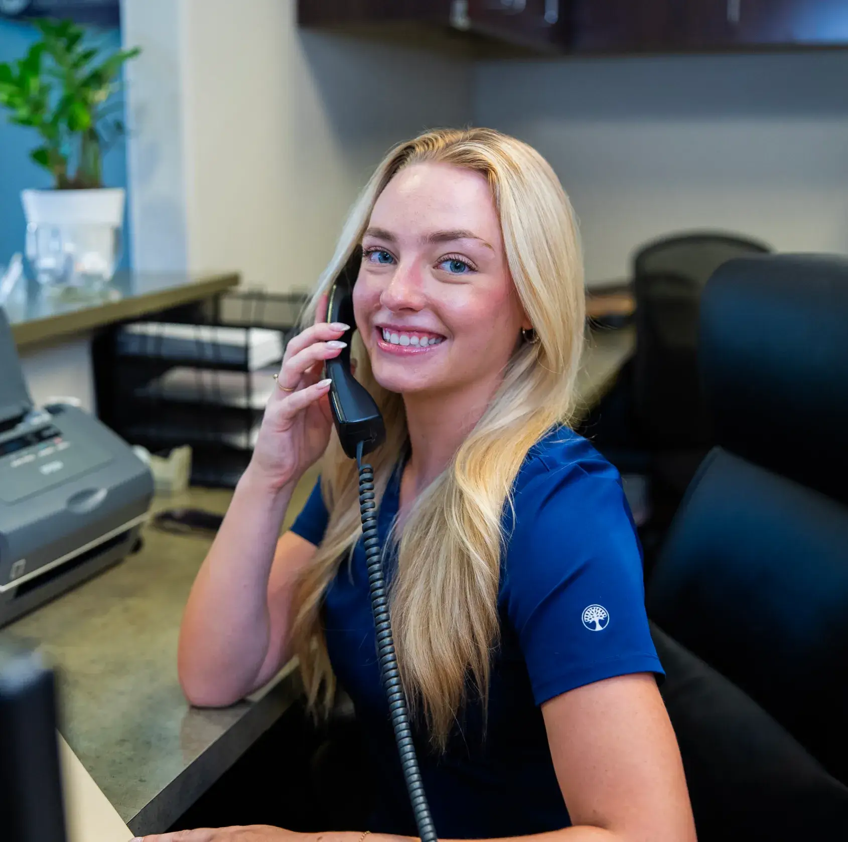 A woman in a blue uniform smiles while talking on the phone at a desk.