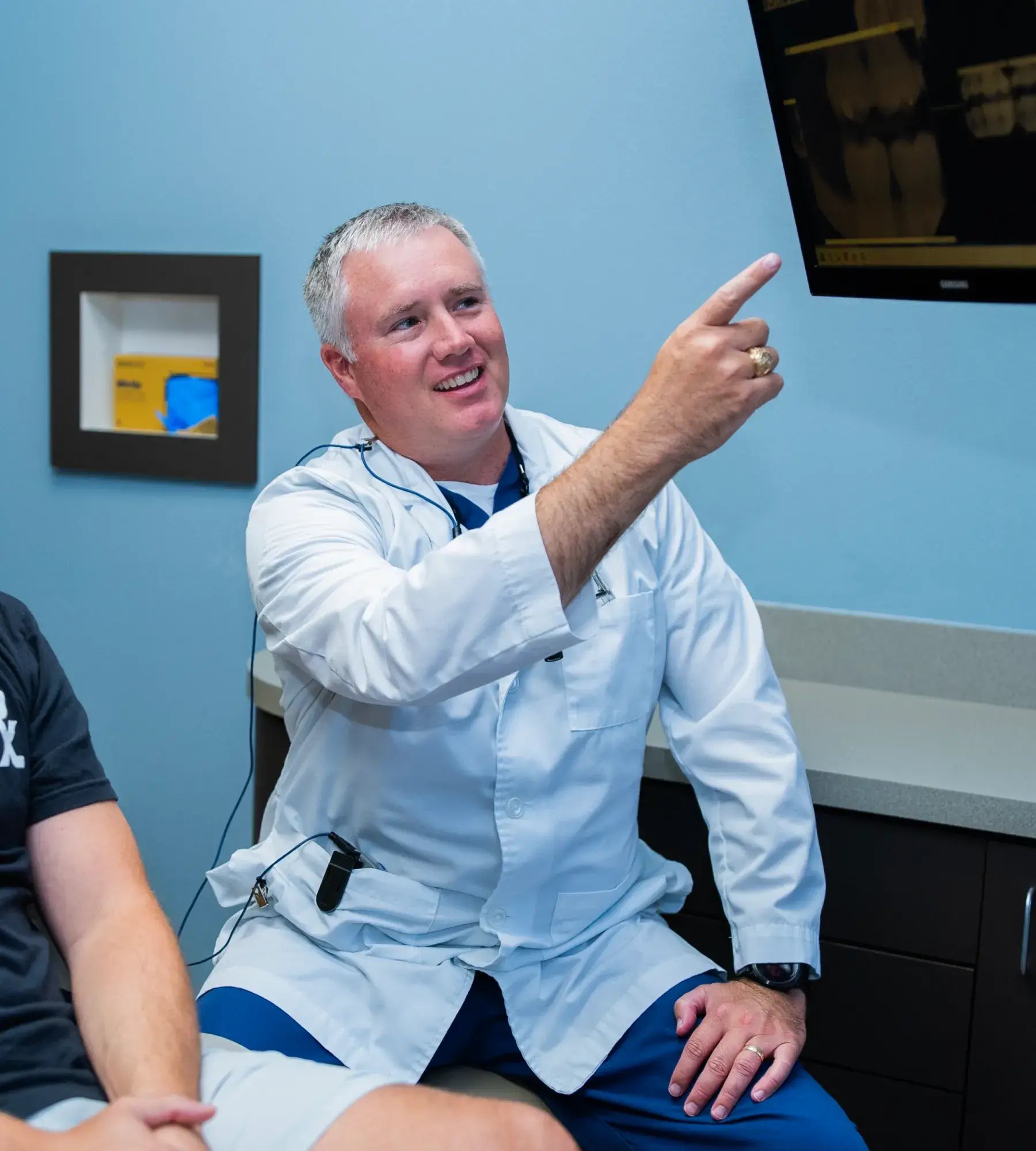 A doctor in a white coat points at an X-ray on a screen while talking to a patient.