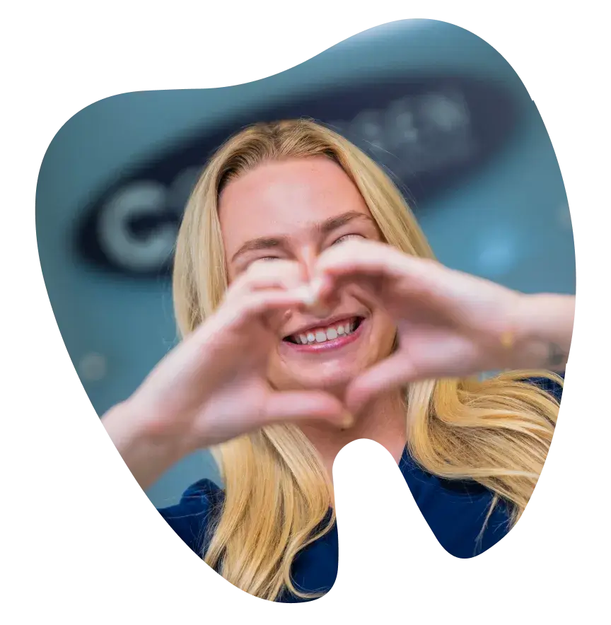 A person with long blonde hair makes a heart shape with their hands and smiles.