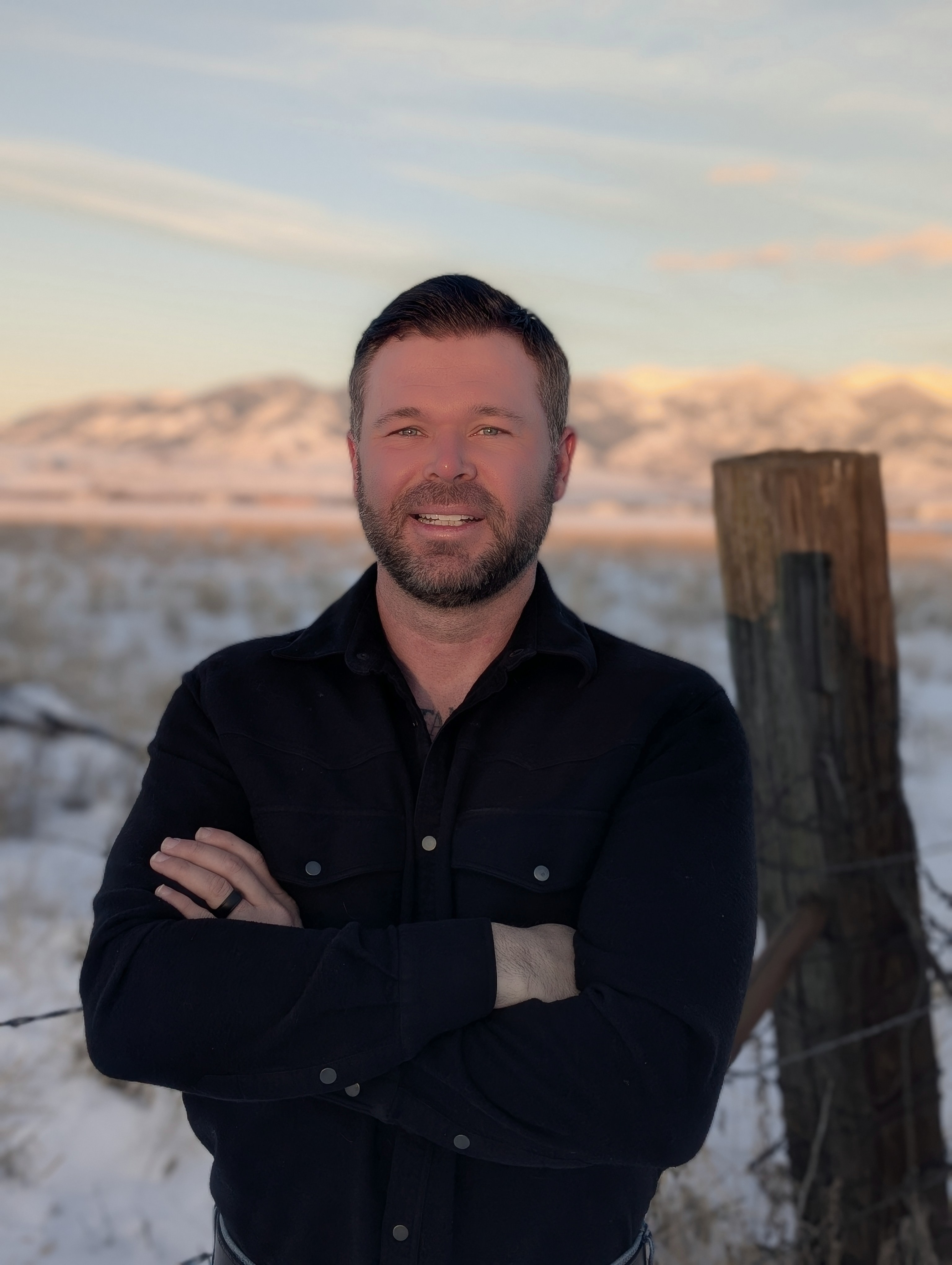 Trent McMurtrey: Man in a black button-up shirt standing in a snowy rural field at sunset, with mountains in the background.