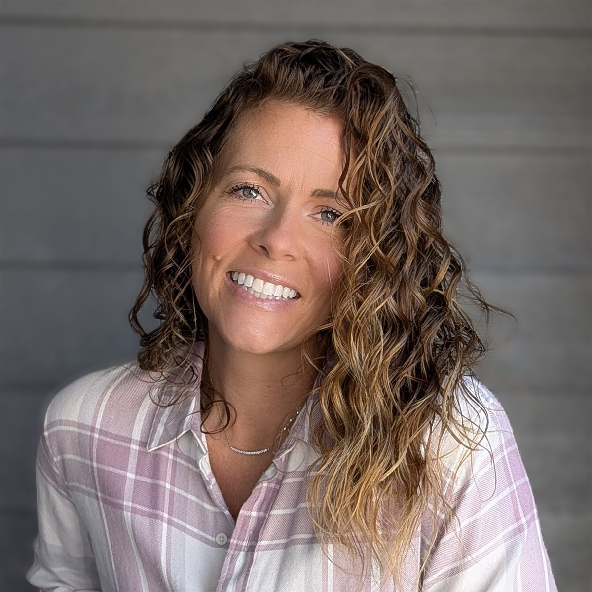 Faith McMurtrey: Woman with wavy hair wearing a light plaid shirt, smiling in front of a neutral wood panel background.