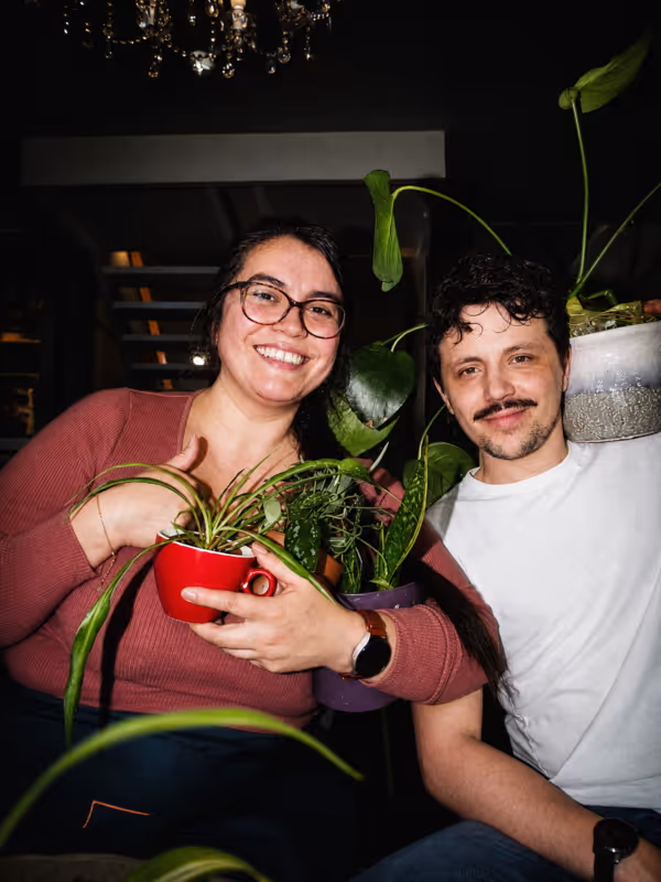 Twee glimlachende mensen zitten dicht bij elkaar binnenshuis, elk een kamerplant vasthoudend in potten.