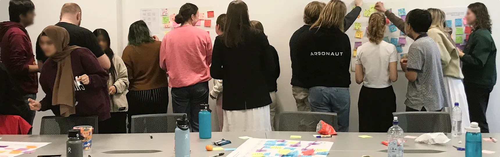 Large group of people, standing in almost two different groups, interacting with post it notes on the wall as the converse.
