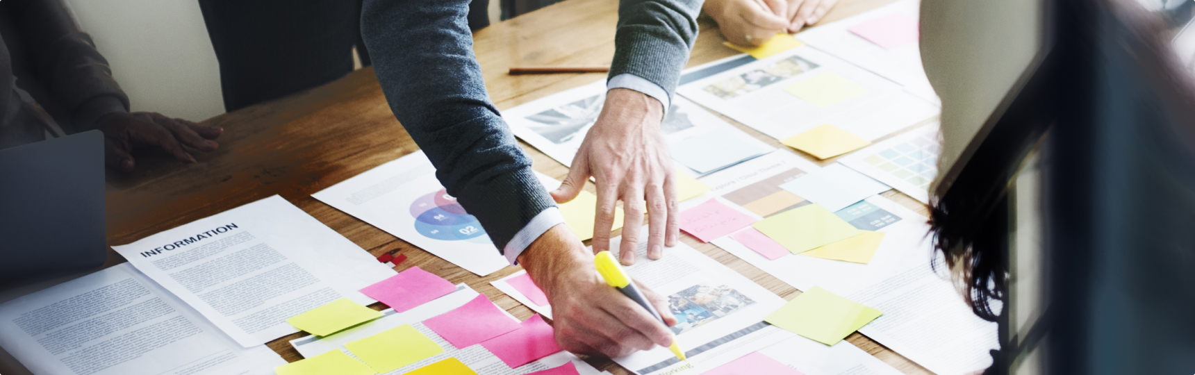 Pages laid out across a wooden table, with multiple people's hands in view. One person reaching over with a highlighter.