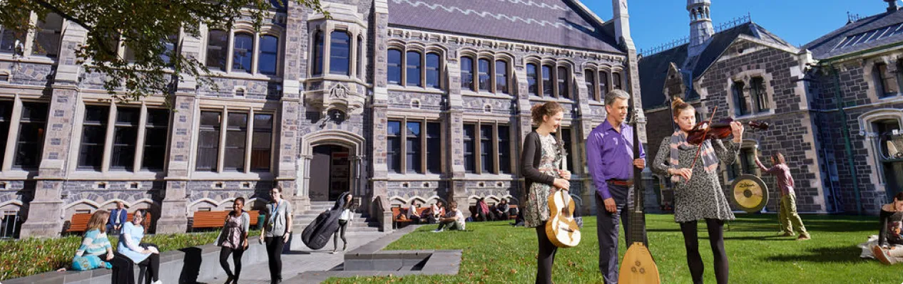Different groups of students standing together on the grass with instruments, while other students are walking on a pathway to and from the building in the background. 