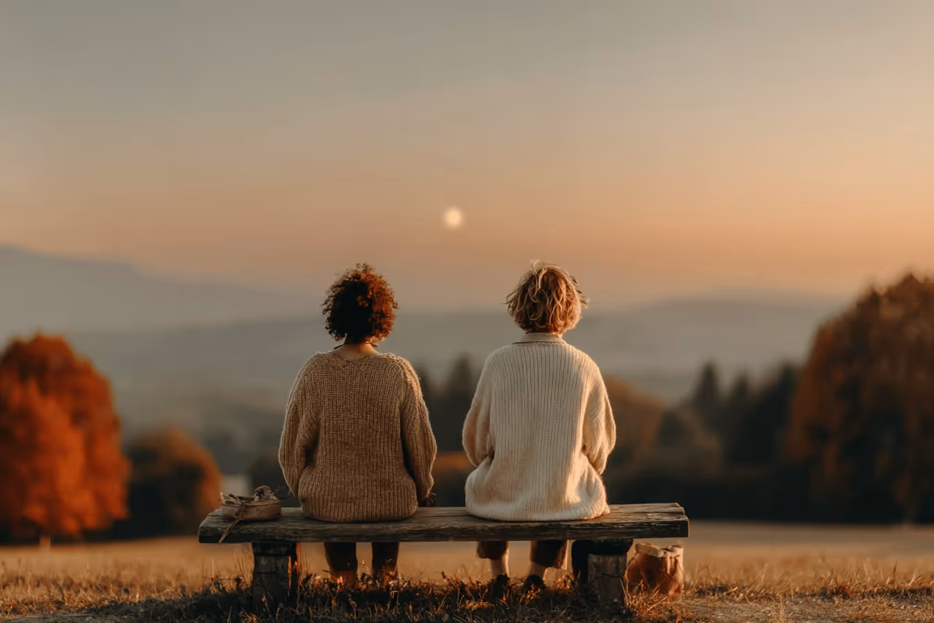 Two women sit on a bench, enjoying a vibrant sunset that casts warm colors across the sky.