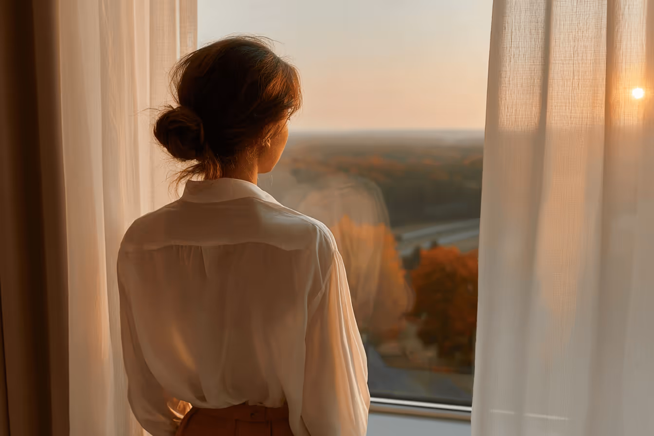A woman looks out a window, captivated by the warm colors of the sunset illuminating the evening sky.