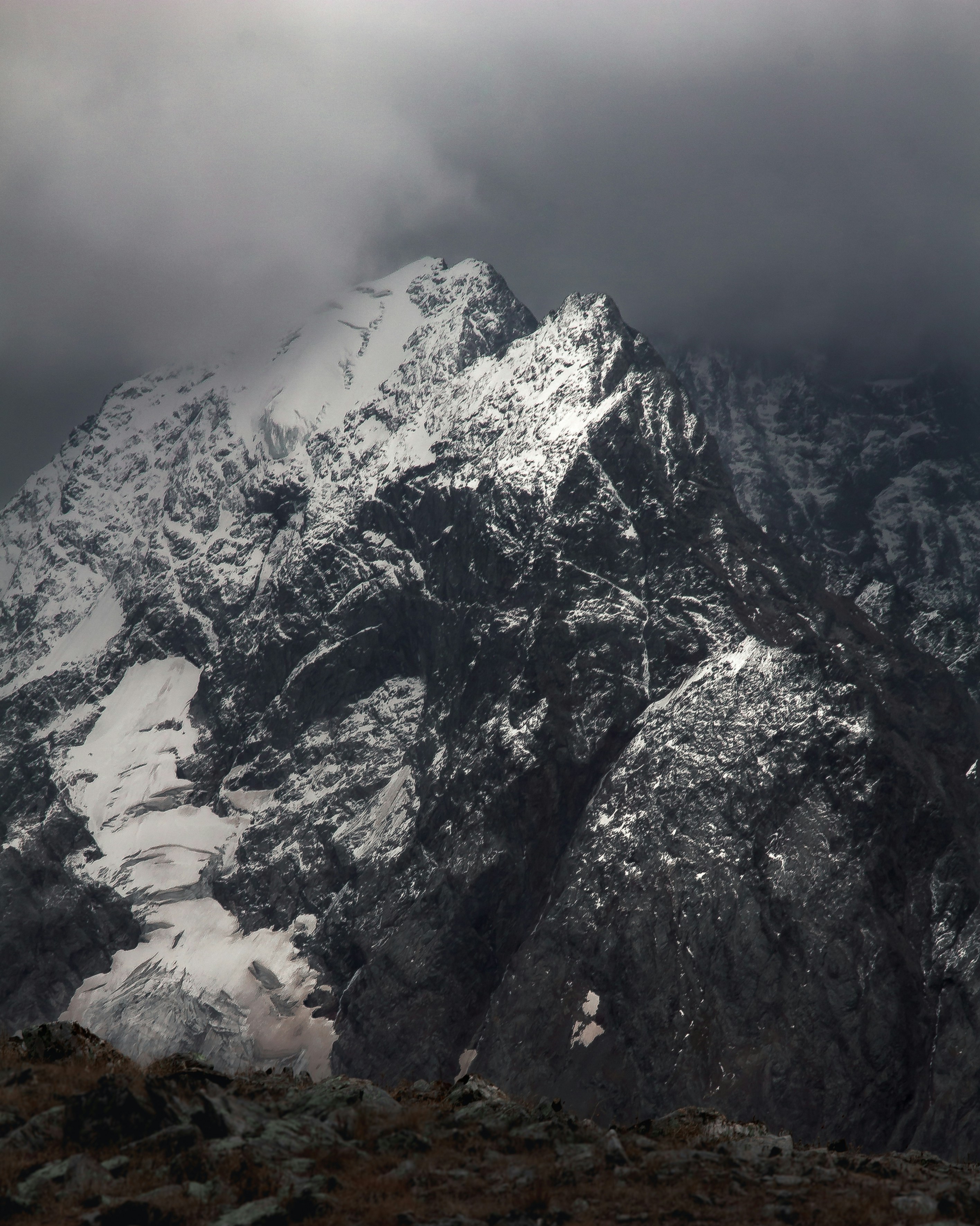 Dramatic snow-covered mountain peak surrounded by dark storm clouds, with rocky terrain in the foreground.
