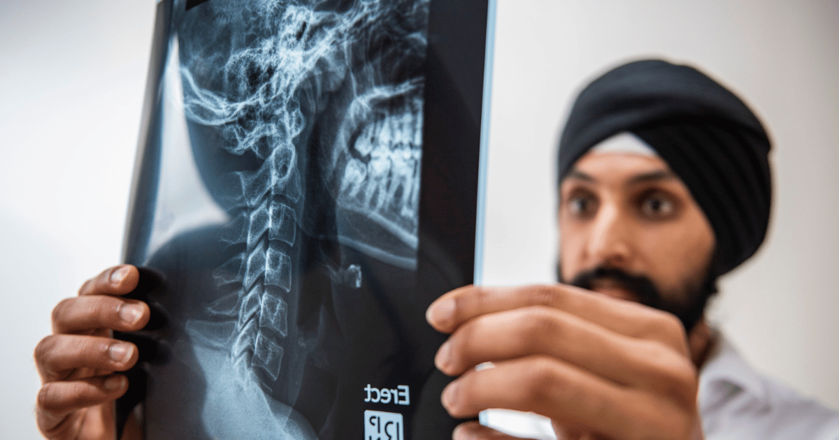 Dr Balraj, a chiropractor wearing a black turban, closely examines a cervical spine X-ray held in front of him, highlighting spinal health assessment in a clinical setting.
