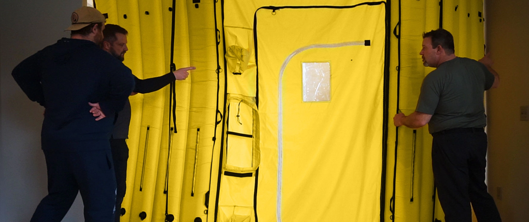 Three men examining and discussing a large yellow inflatable or collapsible structure inside a room.