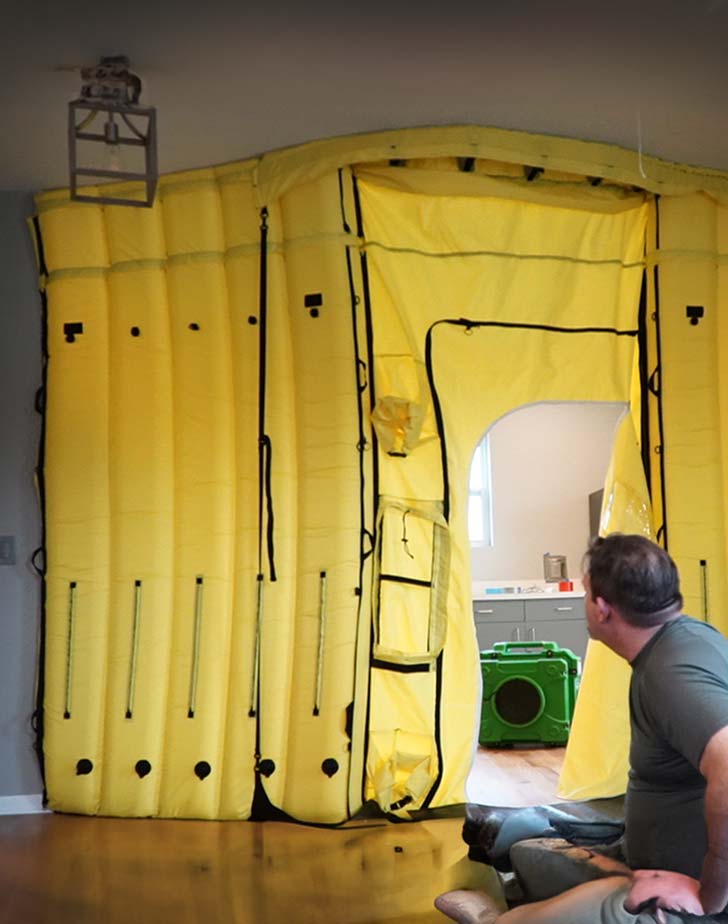 Man sitting on floor looking at a large yellow inflatable containment enclosure with an open doorway inside a room.