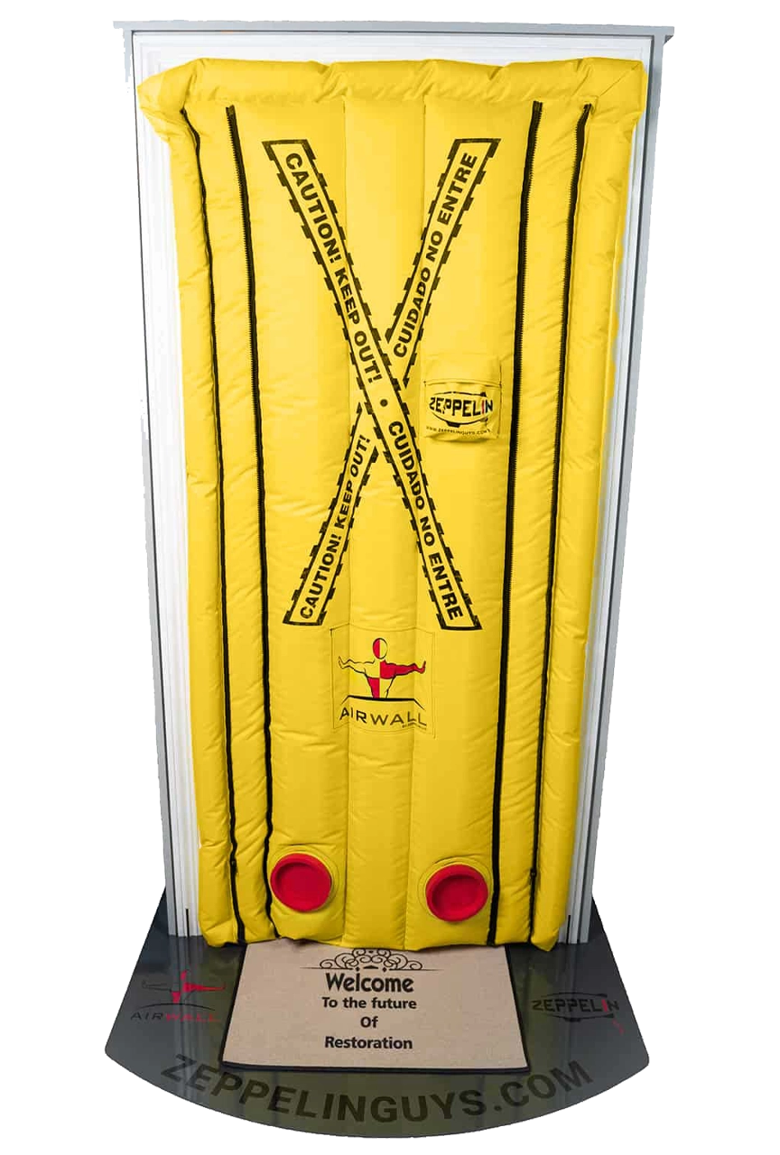Yellow inflatable door seal with black 'Caution! Keep Out!' and 'Cuidado No Entre' tape markings, red circular valves, and black zippers on each side, installed in a white door frame over a mat reading 'Welcome to the future of restoration'.