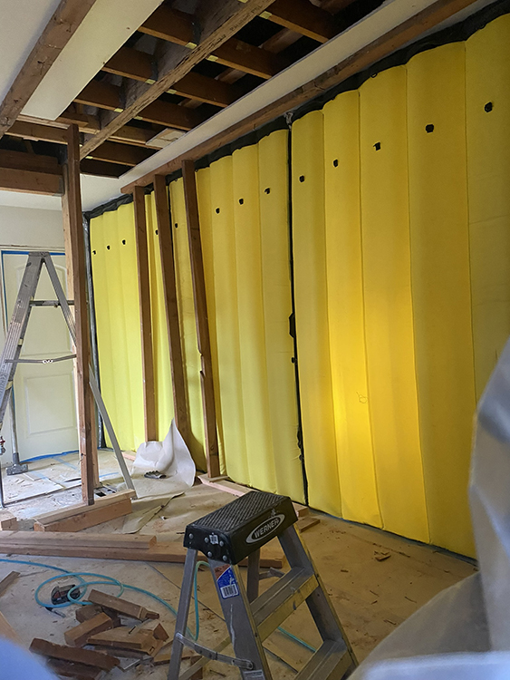 Indoor construction site with exposed wood framing, yellow inflatable air barrier on the wall, and a Werner step ladder in the foreground.