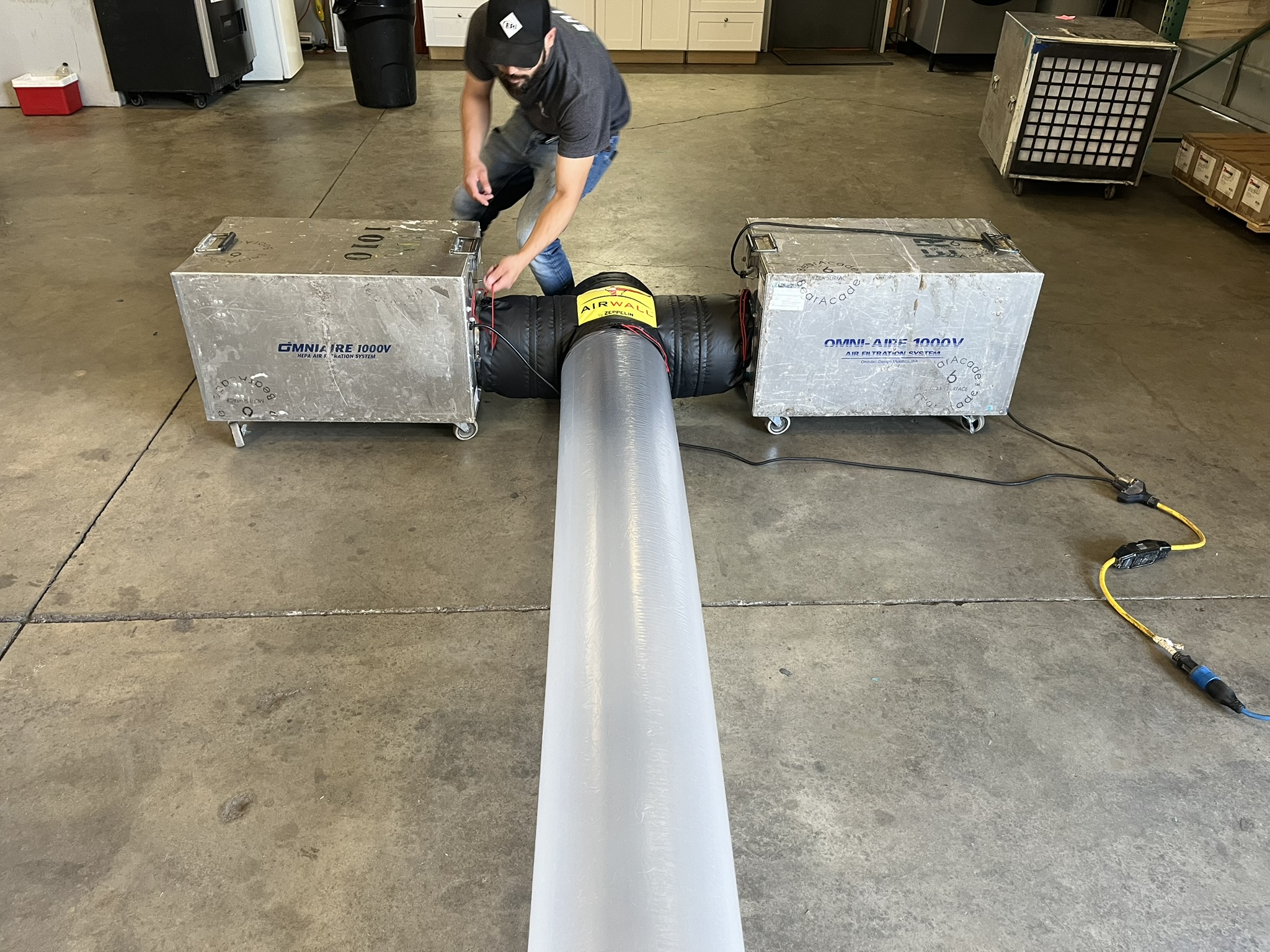 A man in a warehouse adjusting wires connected to two metal air filtration units joined by black and white flexible ducting labeled Airwall Zeppelin.