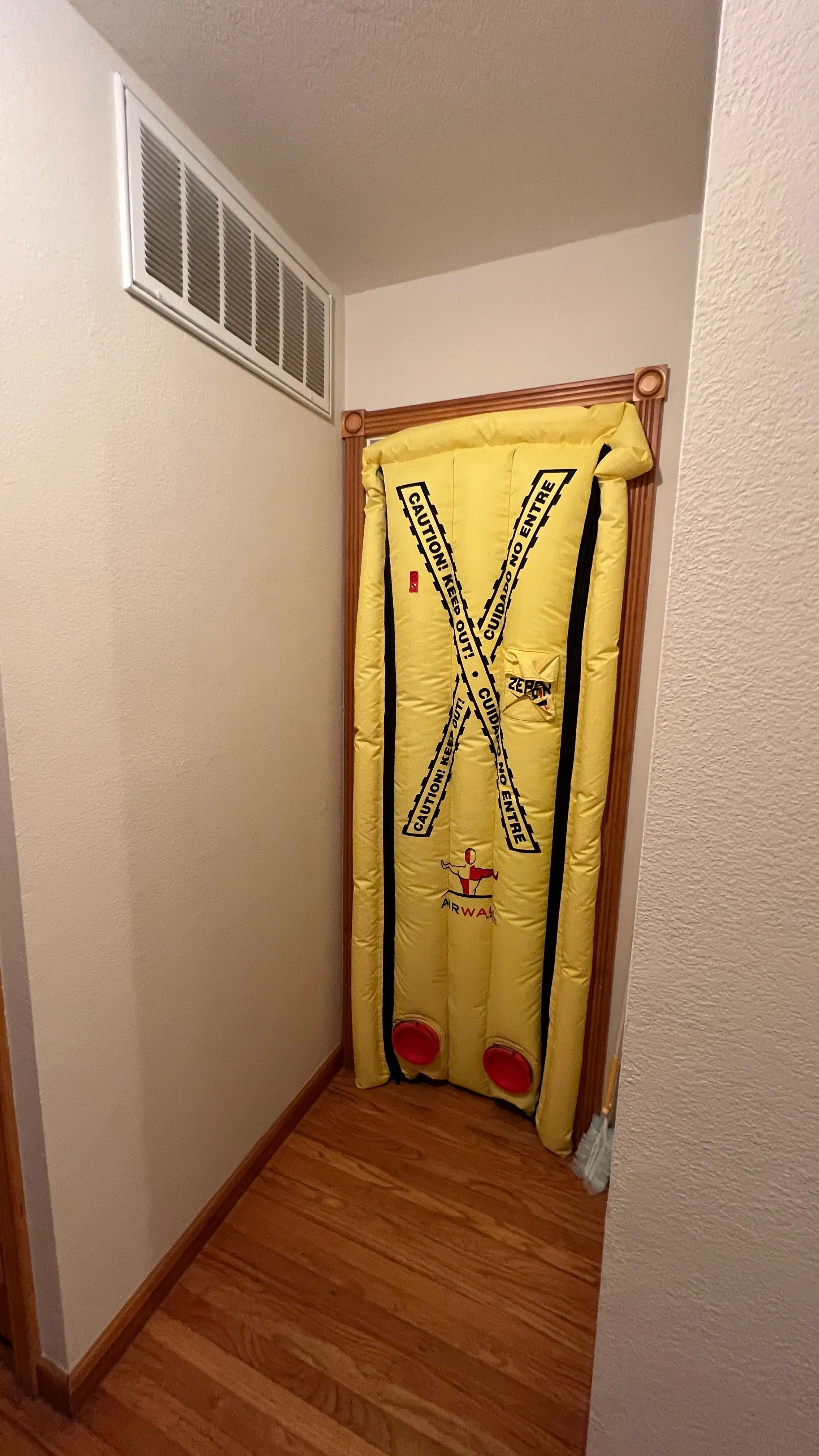 Yellow inflatable air barrier with caution tape design blocking a doorway in a hallway with wooden floor and beige walls.