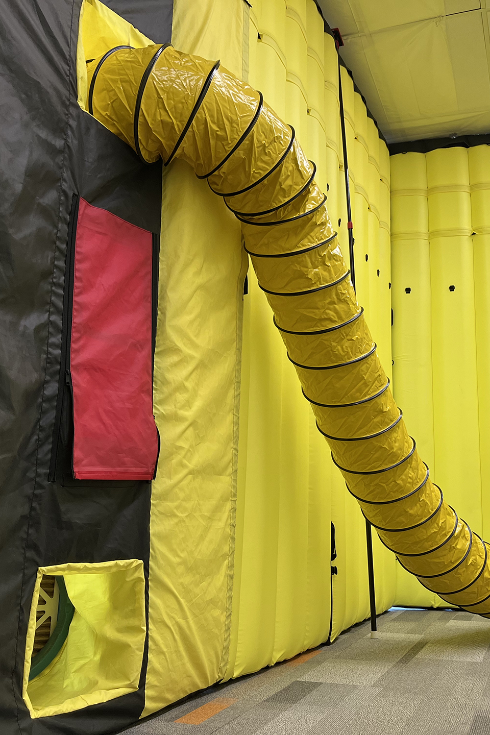 Yellow flexible ventilation duct extending from a yellow inflatable wall with red and black fabric panels.