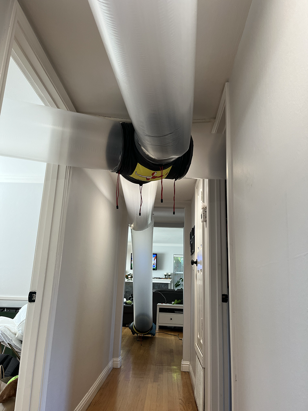 Two large connected flexible ventilation ducts running through a hallway with wooden floor and white walls.