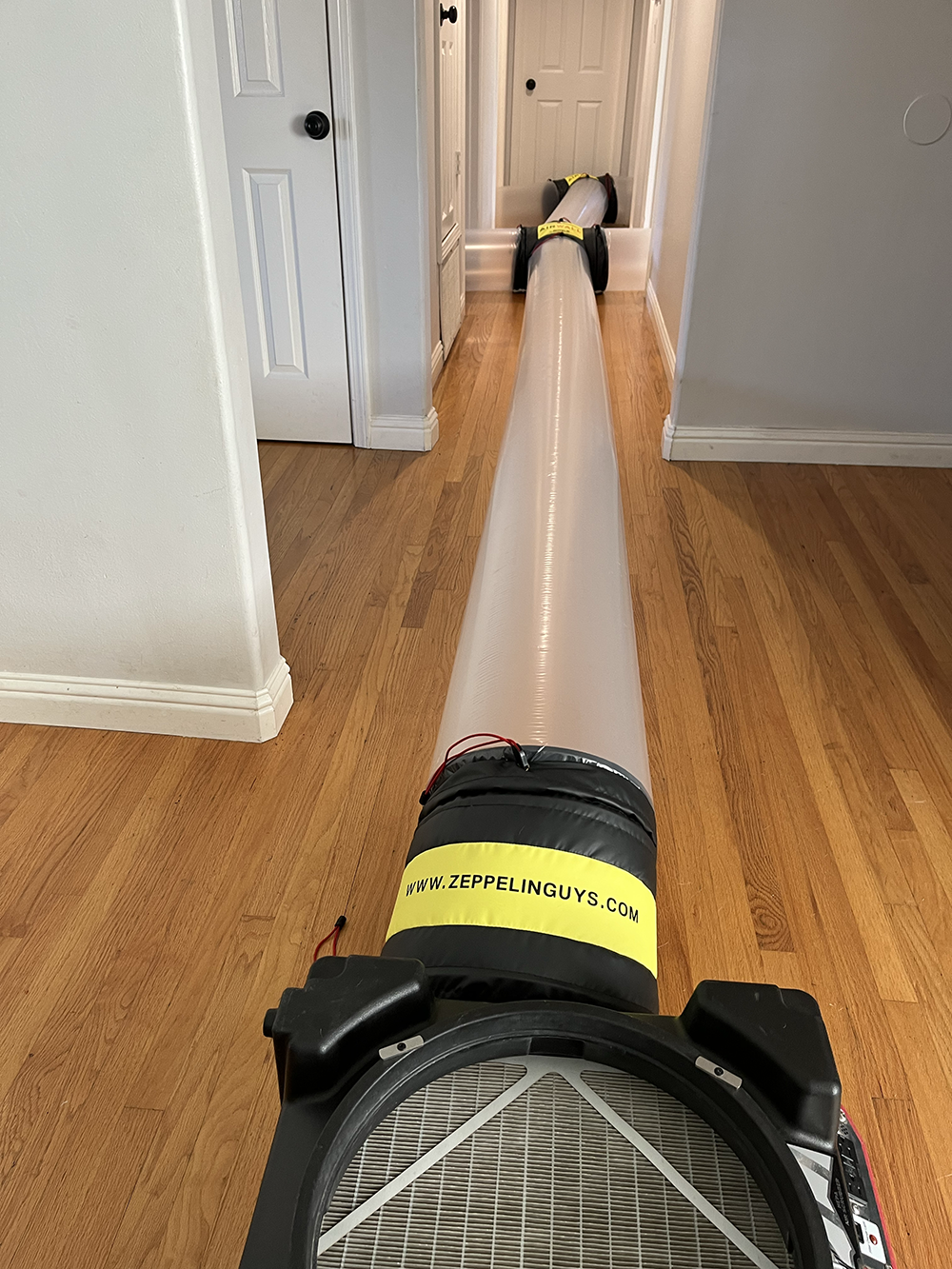 Long cylindrical air duct with a black fan unit at the near end on a wooden floor in a hallway.